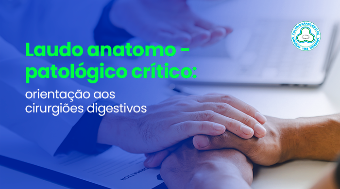 Médico segurando as mãos de um paciente durante uma consulta, em gesto de acolhimento, enquanto conversa em um consultório com prontuário e computador sobre a mesa.