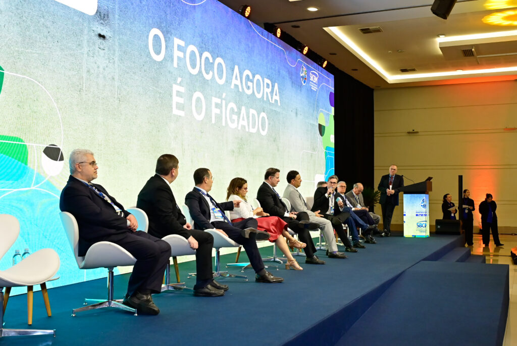 pessoas sentadas num palco numa roda de debate, escrito no telão "o foco é o fígado"