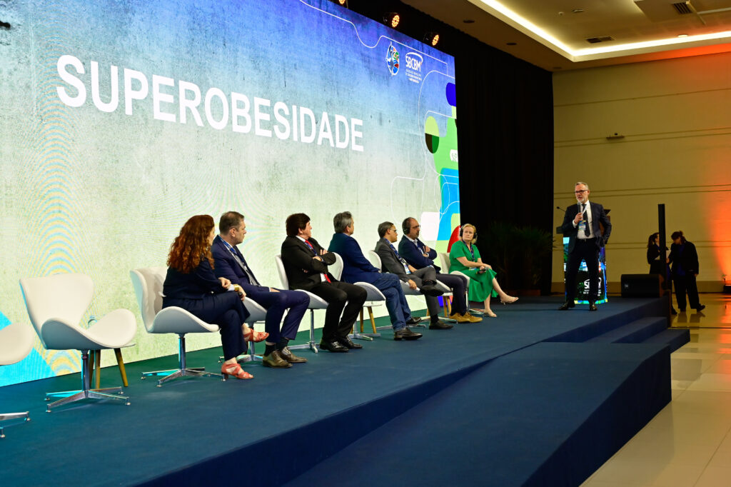 pessoas sentadas num palco numa roda de debate, escrito no telão "superobesidade"